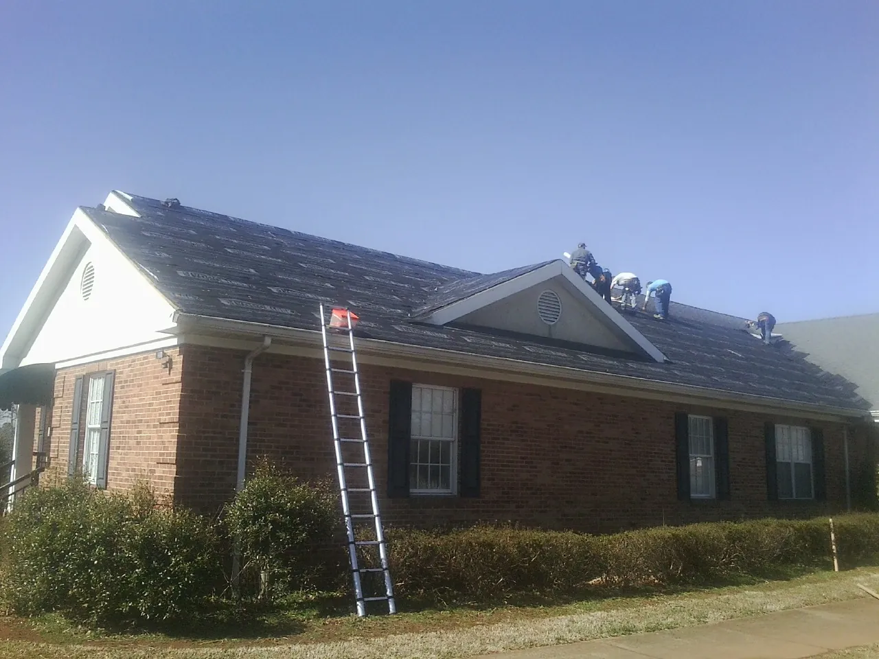 Crew tearing shingles off a brick ranch roof with a ladder set — Metal Roof Repair work by our crew in De Pere