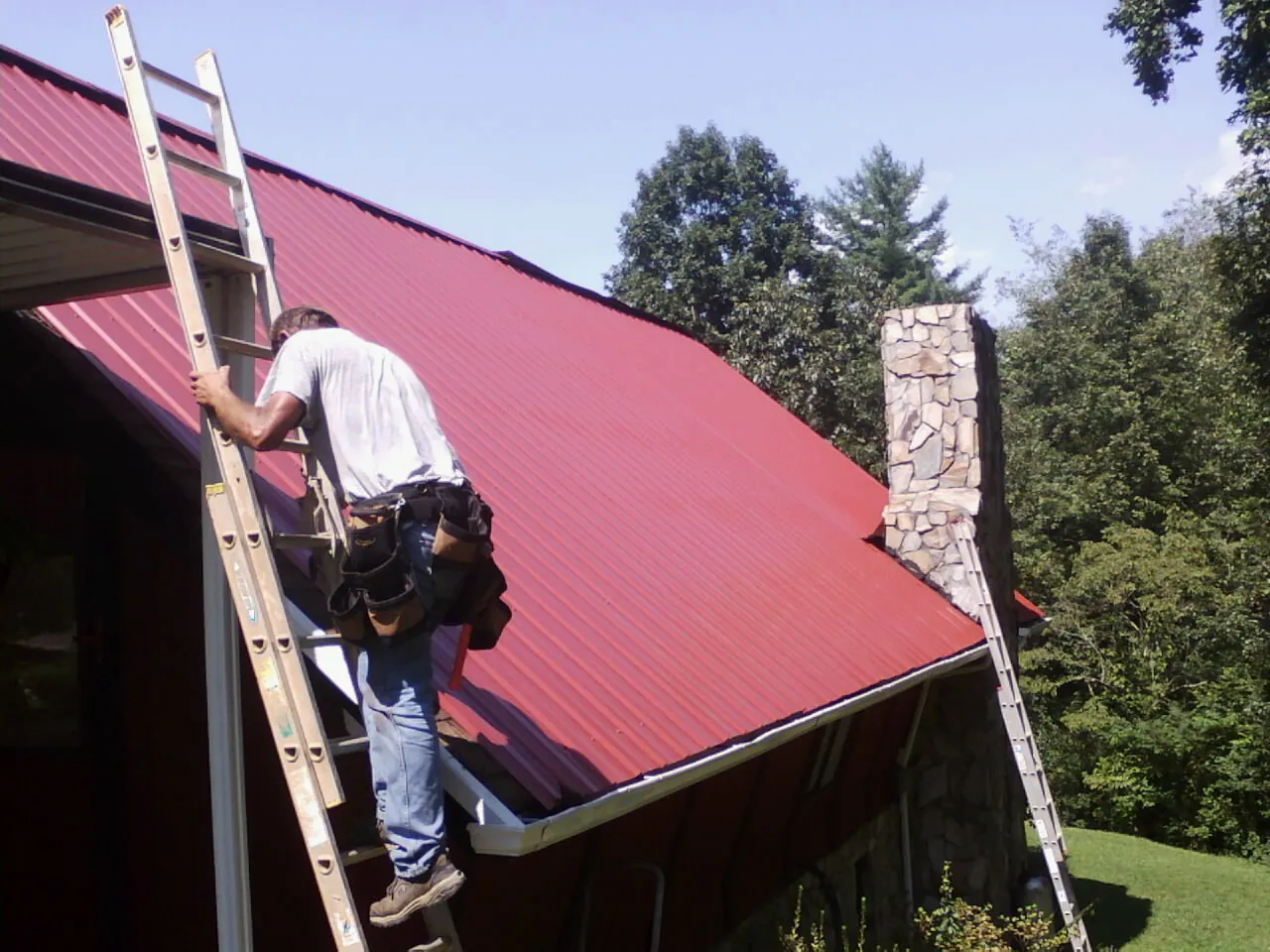 Roofer on a ladder beside a red metal roof and stone chimney — Soffit & Fascia Repair by Roofing Brothers in De Pere